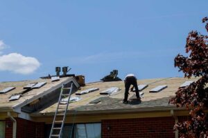 A roof worker installing a small peaked structure behind a chimney to divert water on an asphalt shingle roof.