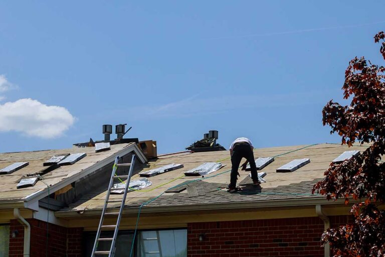 A roof worker installing a small peaked structure behind a chimney to divert water on an asphalt shingle roof.
