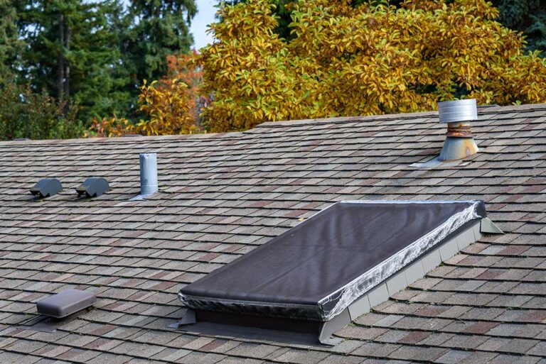 Roof worker inspecting metal flashing around a plumbing vent on a shingled roof.